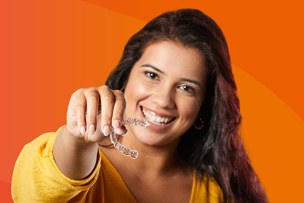 woman in yellow holding out a SureSmile clear aligner Transparent SureSmile aligner being held by a patient, offering a discreet and comfortable alternative to traditional braces.